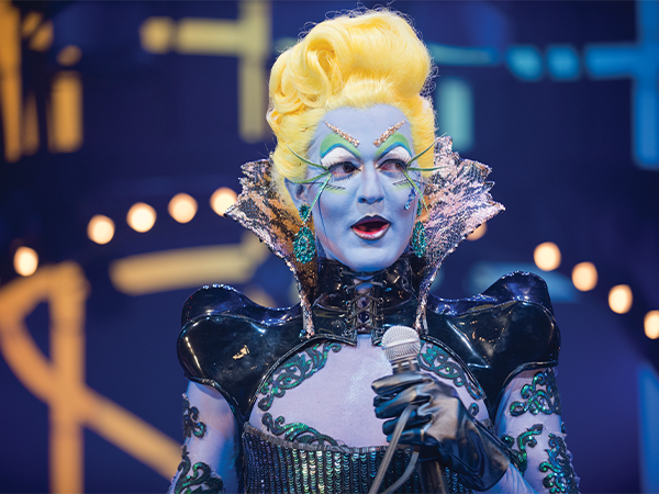 A person dressed in elaborate costuming and blue makeup and yellow hair speaks on the stage during a theatre production.